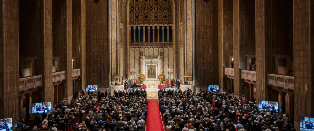 A large congregation gathers for a special event in a majestic sanctuary with soaring ceilings and a central aisle adorned with a red carpet.