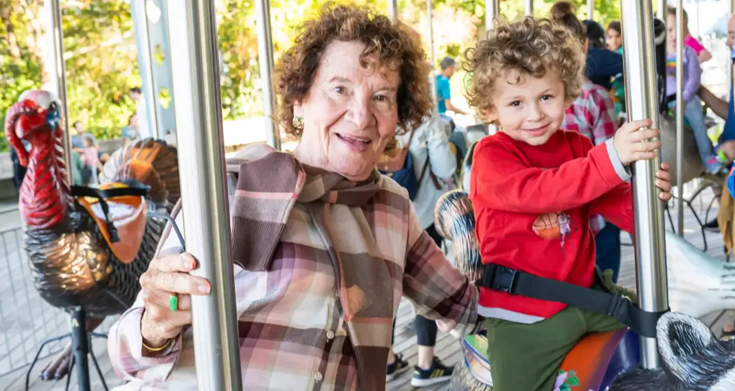 Smiling elderly woman and young boy with curly hair ride a carousel together, sharing joyful moments at a Temple Emanu-El event.
