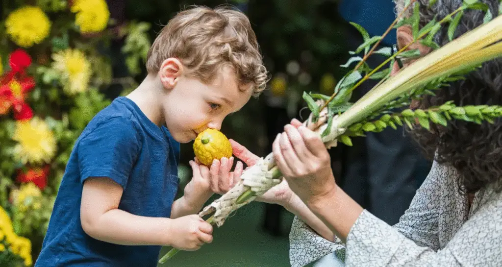 A child enjoys the scent of an etrog as an adult holds a lulav, both surrounded by colorful flowers during Sukkot at Temple Emanu-El.