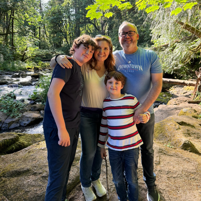 A family of four smiles together beside a stream in a sunlit, green forest, enjoying time outdoors as a community.