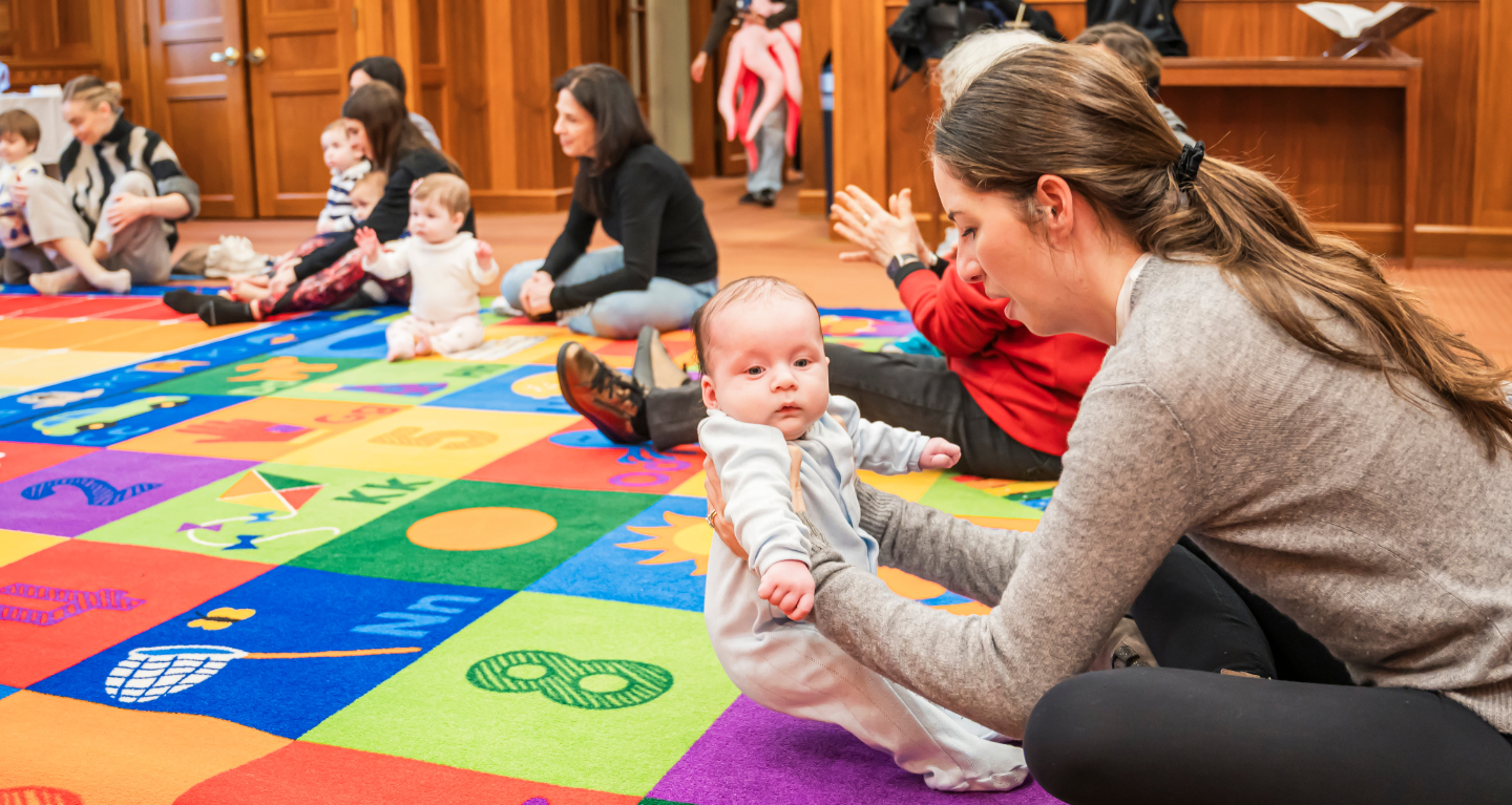 Families with young children gather on a colorful play mat in a wood-paneled room at Temple Emanu-El, enjoying group activities.