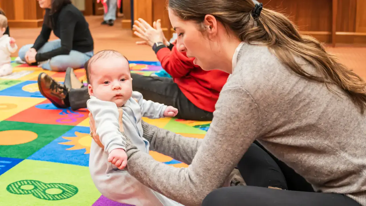 A woman holds a baby upright on a colorful play mat during a group gathering at Temple Emanu-El, with others in the background.