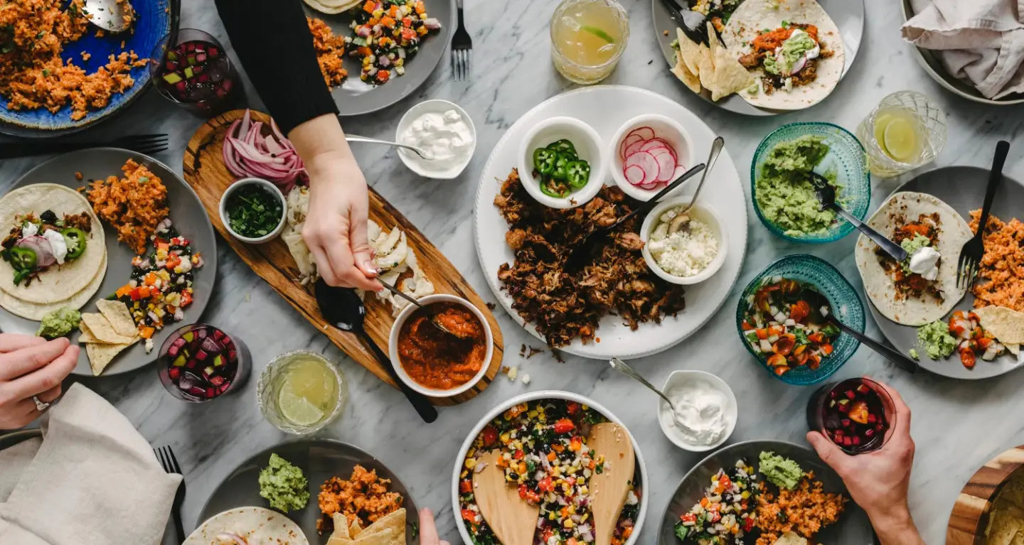 Hands reaching for tacos, rice, guacamole, and salsa on a table set for a communal meal with beverages at Temple Emanu-El.