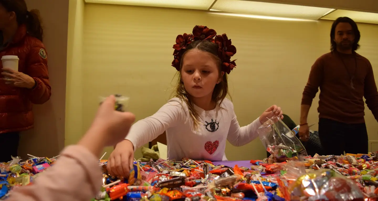 Young girl with a flower headband reaches for treats at a table covered with assorted snacks during a Temple Emanu-El event.