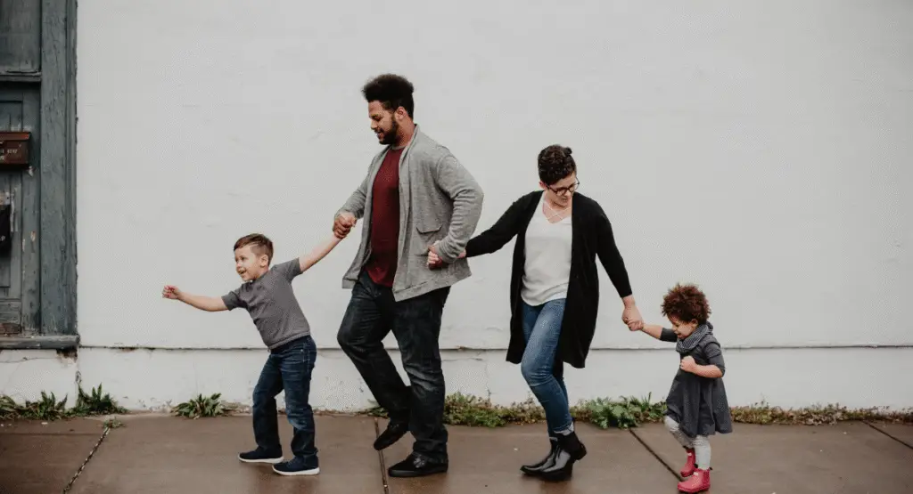 Four individuals holding hands, walking together along a sidewalk in front of a white building, symbolizing community and togetherness.