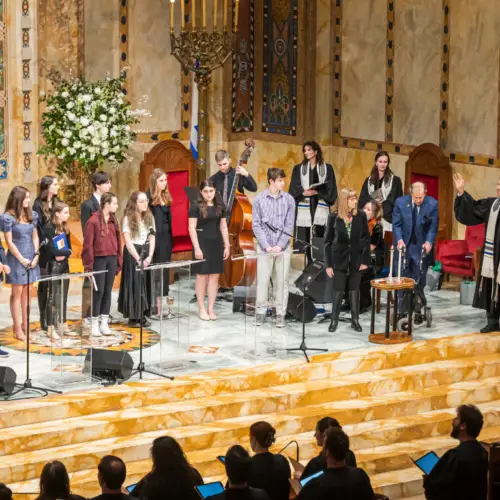 Congregants and clergy in robes stand on the bimah at Temple Emanu-El during a service, with the congregation seated below.
