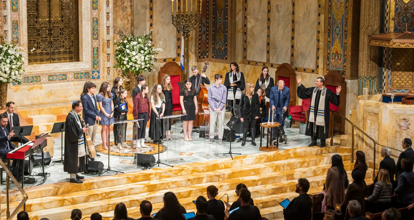 Congregants and clergy in robes stand on the bimah at Temple Emanu-El during a service, with the congregation seated below.