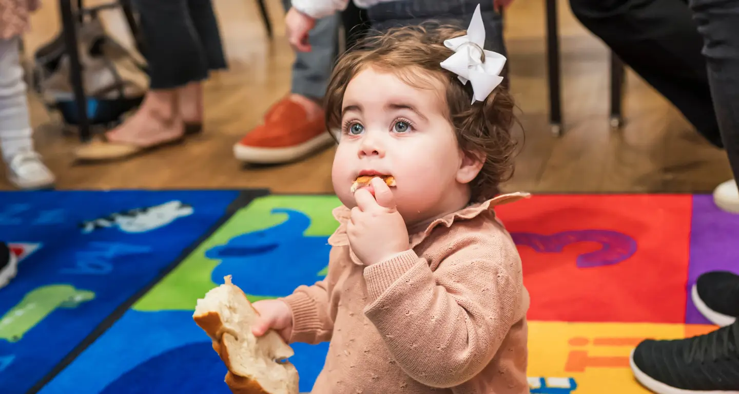 Toddler with a white bow enjoys bread while sitting on a colorful mat during an indoor gathering at Temple Emanu-El.