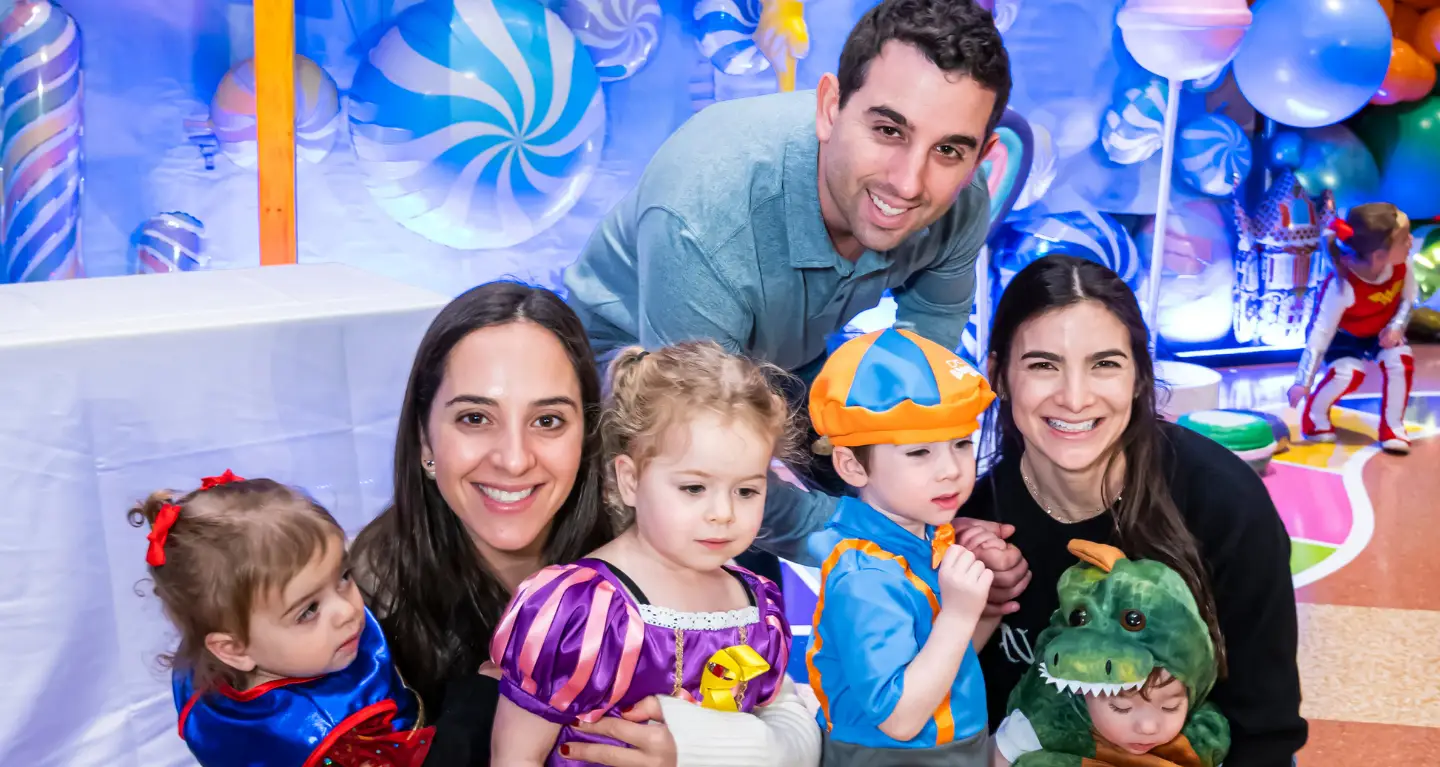 Three adults and three young children in costumes pose and smile at a vibrant, festive celebration at Temple Emanu-El.