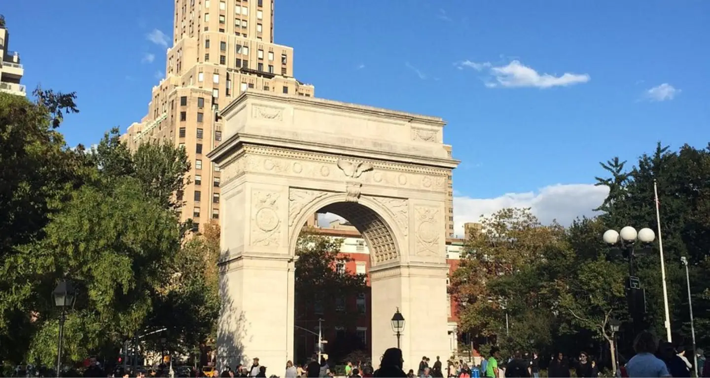 Washington Square Arch in New York City with people gathered nearby; city buildings and vibrant urban life form the backdrop.