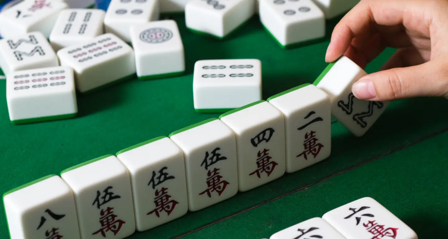 A hand arranges Mahjong tiles on a green table at a community gathering, with various tiles spread around.