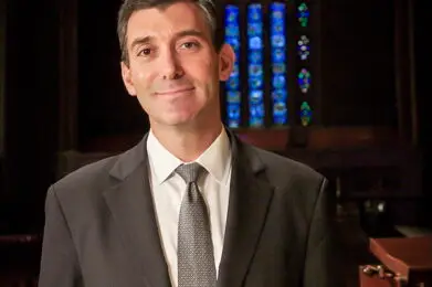 Rabbi Joshua Davidson in a suit standing inside Temple Emanu-El, with a large, colorful stained glass window behind him.