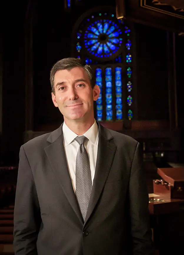 Rabbi Joshua Davidson in a suit standing inside Temple Emanu-El, with a large, colorful stained glass window behind him.
