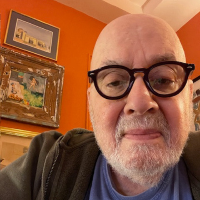 Bald man with glasses taking a selfie in an orange-walled room , with framed art on the wall behind him.