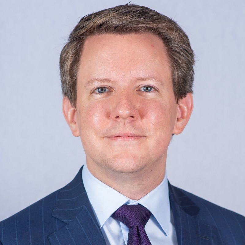 A man wearing a blue pinstripe suit and purple tie smiles warmly at the camera, standing before a neutral light background.