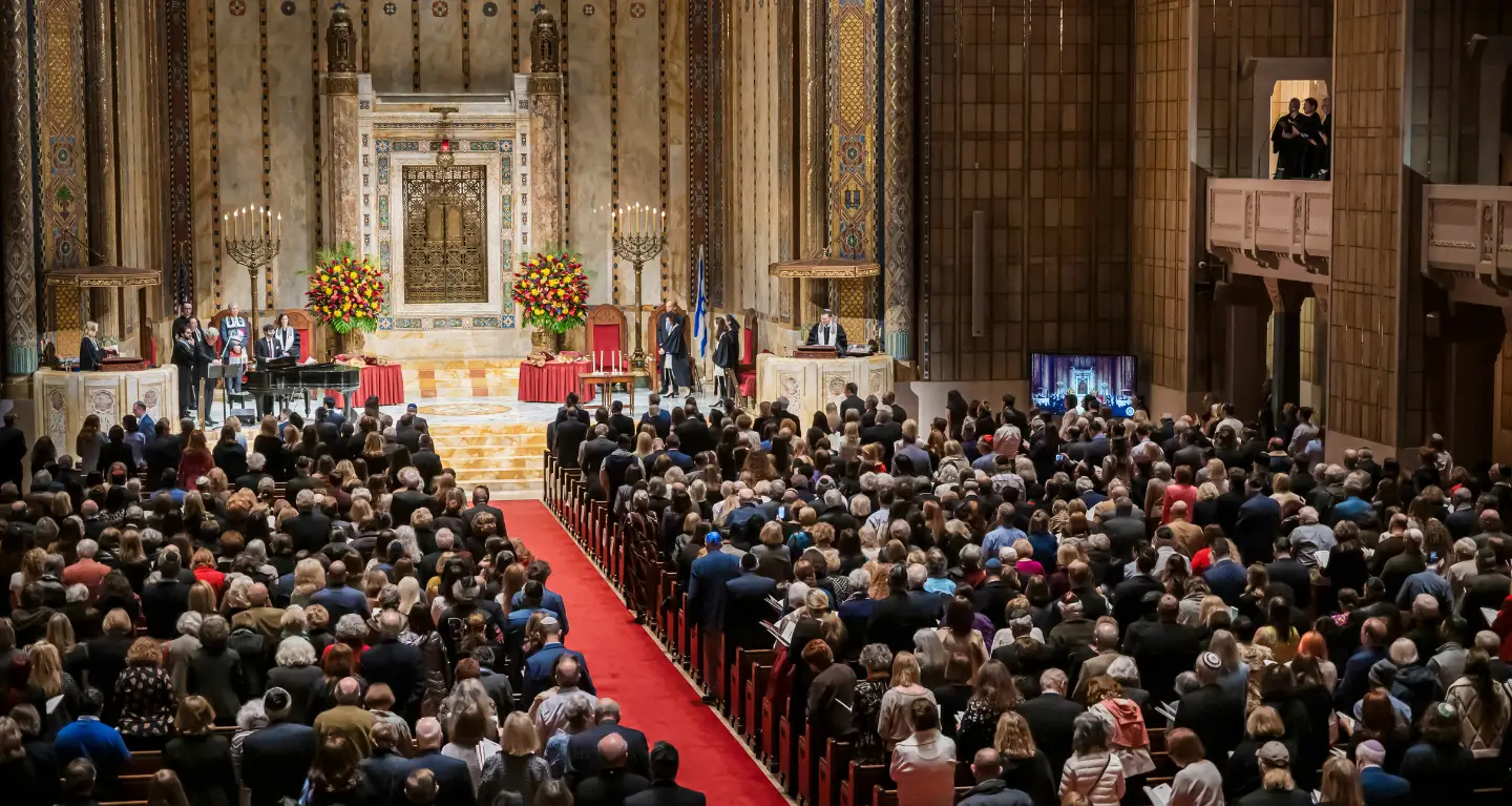 Large congregation seated in grand sanctuary with speakers at the bimah, facing an adorned ark and floral arrangements.