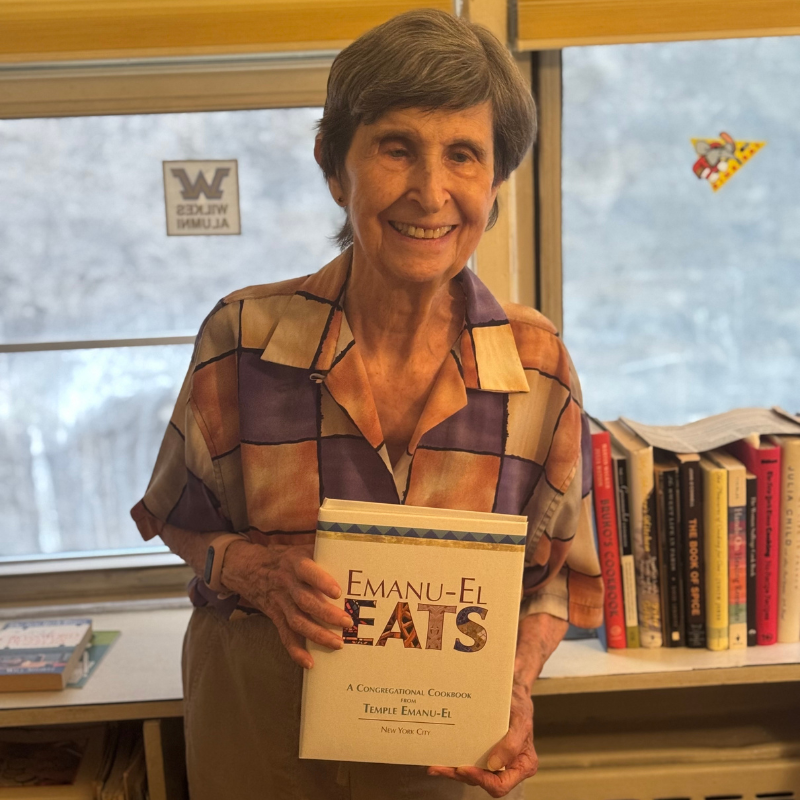 Smiling congregant holding a cookbook titled "Emanu-El Eats" in a warmly lit room with bookshelves and windows.