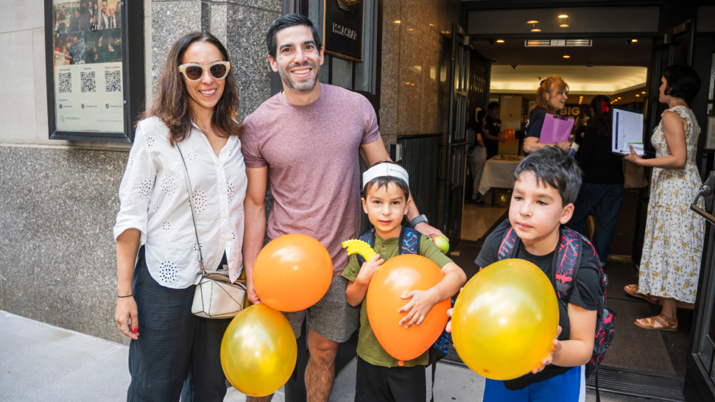 A smiling family with two boys holding orange and yellow balloons stands outside Temple Emanu-El in New York City.