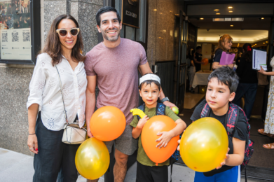 A smiling family with two boys holding orange and yellow balloons stands outside Temple Emanu-El in New York City.