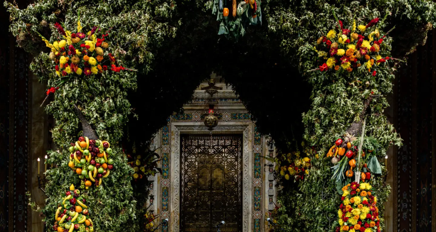 A synagogue entrance framed by leafy branches and colorful floral and fruit arrangements on each side, evoking celebration.