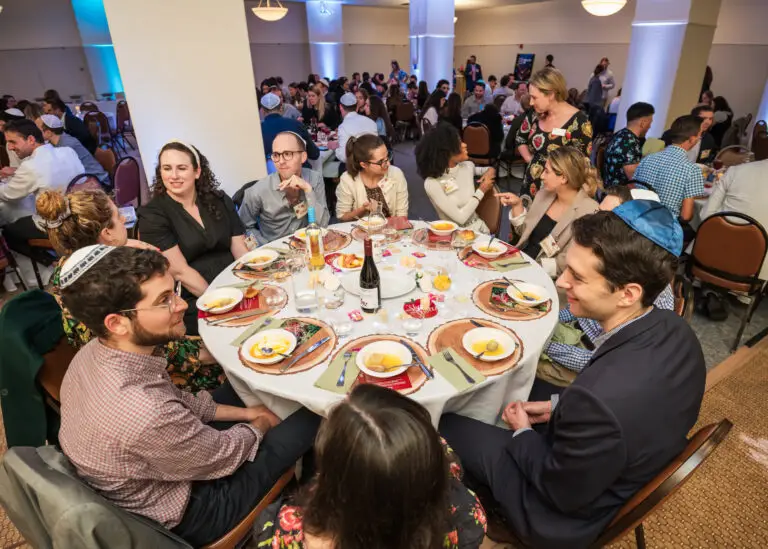 Congregants sit around a round table, sharing food during a lively community event at Temple Emanu-El.