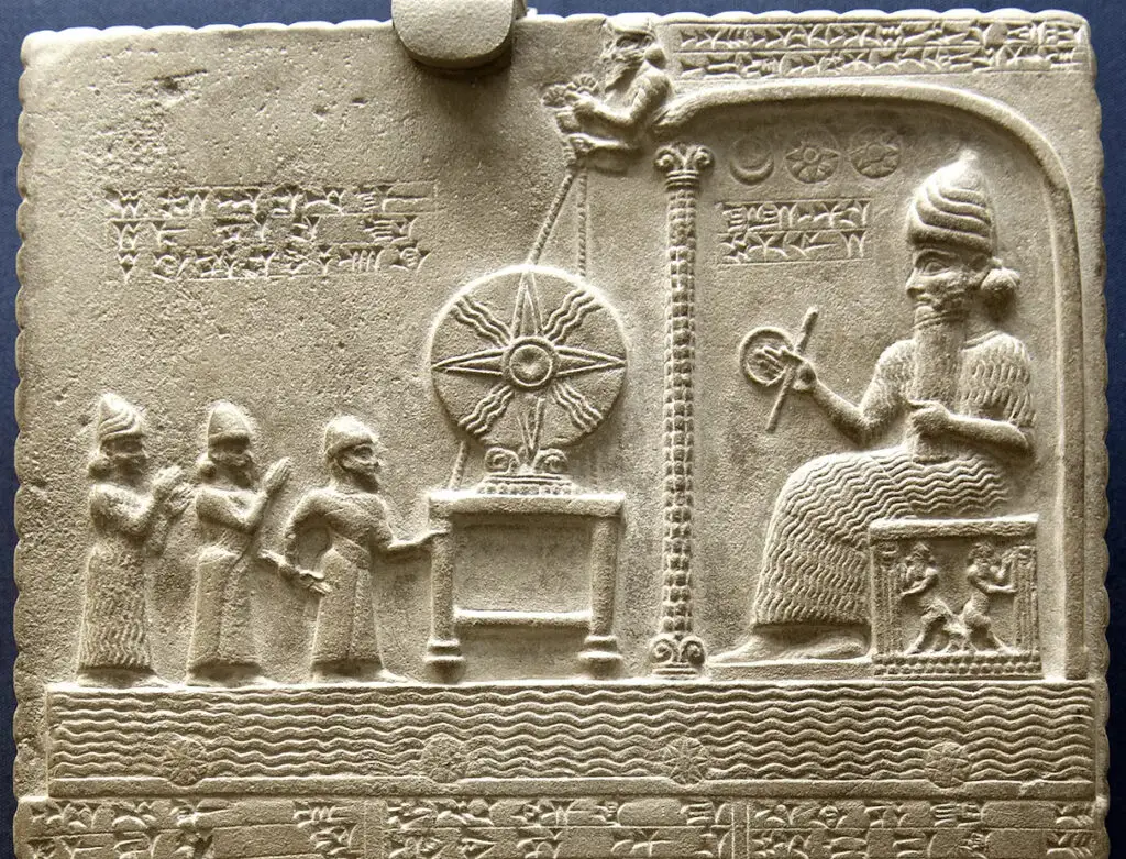 Ancient stone relief with four figures, decorative symbols, cuneiform script, and a seated figure holding a ring and staff.