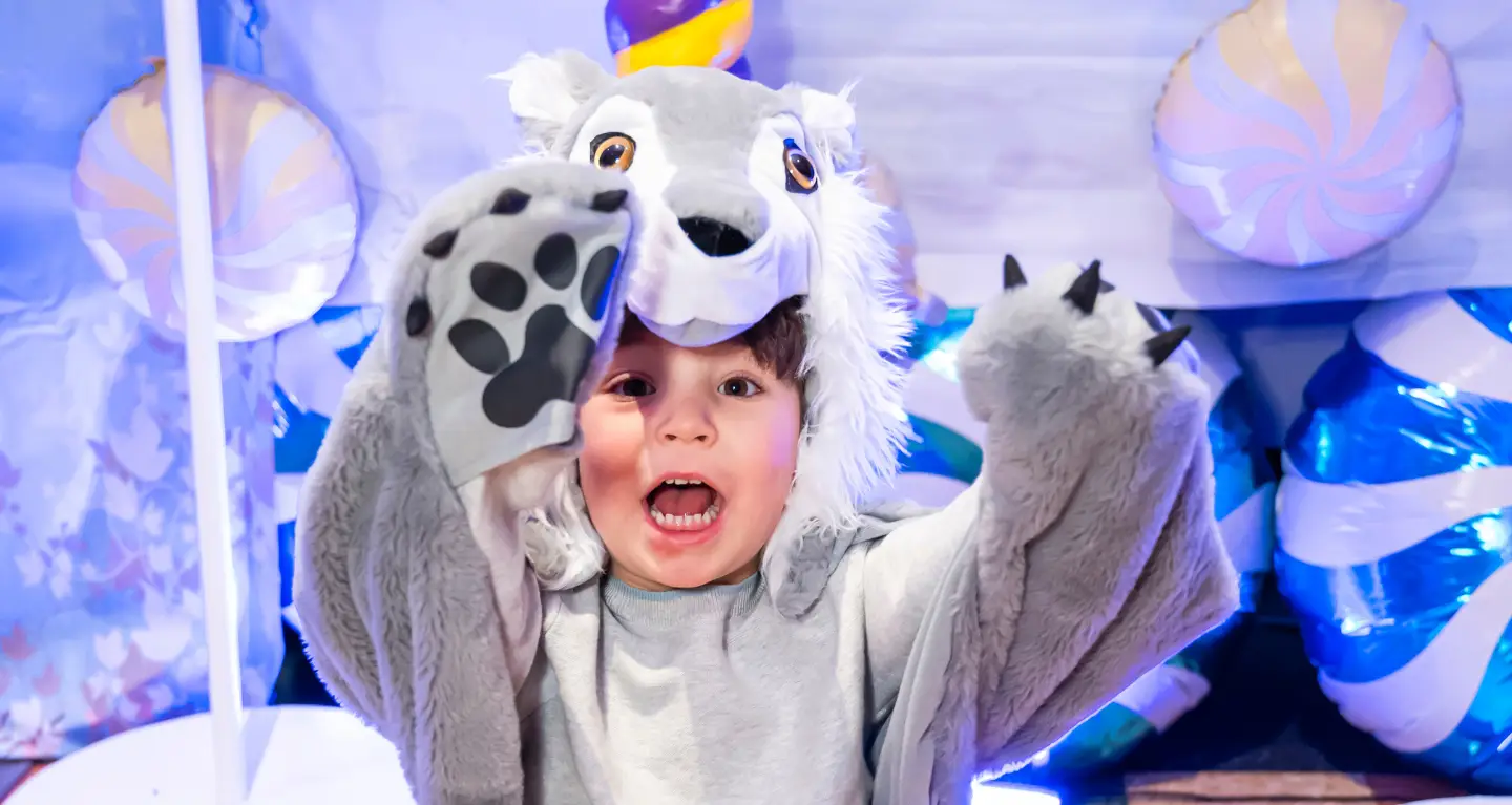 A child in a wolf costume smiles and lifts their paws, surrounded by vibrant, festive decorations at Temple Emanu-El.