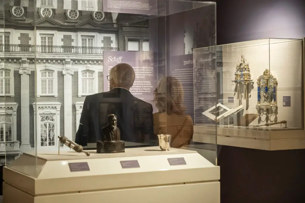 Two visitors look at Jewish historical artifacts in glass cases at Temple Emanu-El, reading informative exhibit plaques.