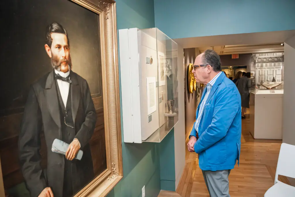 A man in a blue jacket observes an exhibit at Temple Emanu-El beside a large portrait of a bearded man.