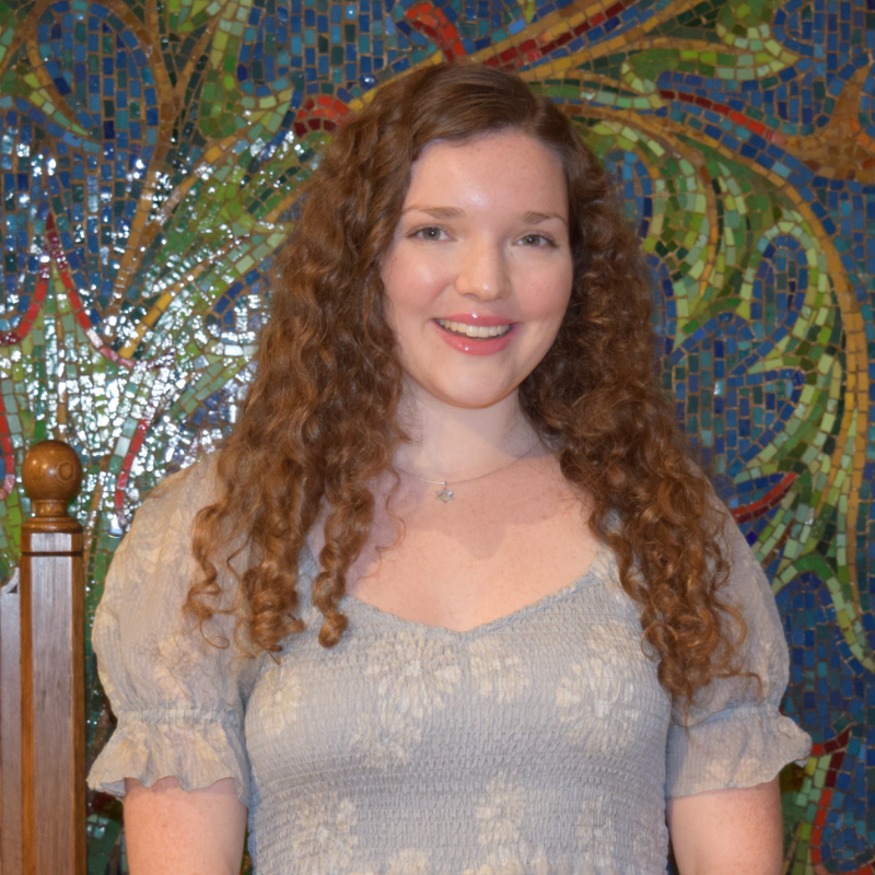 A woman with long curly hair smiles warmly in front of a vibrant mosaic at Temple Emanu-El in New York City.