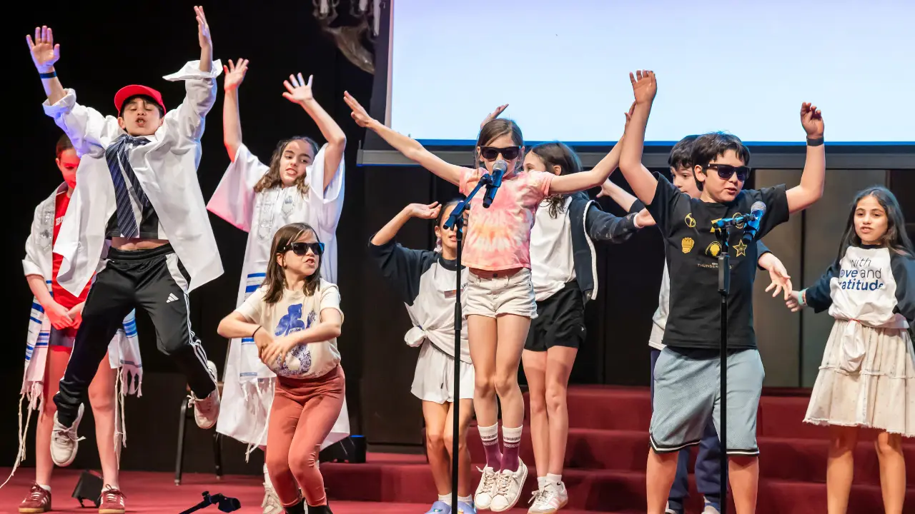 Children on stage at Temple Emanu-El, wearing sunglasses, singing and dancing enthusiastically into microphones during a performance.