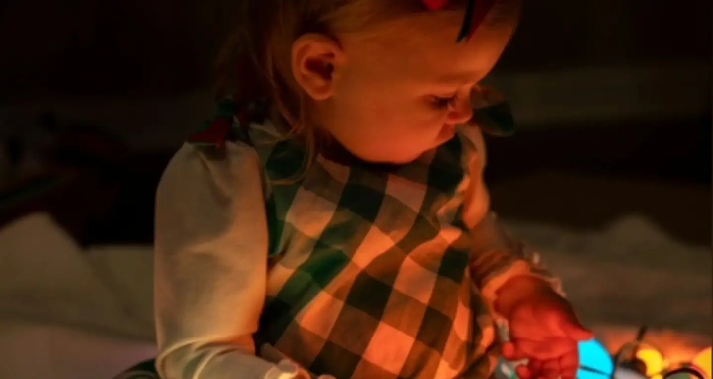 A young child in a green plaid dress gazes at colorful glowing lights in a softly lit room at Temple Emanu-El.