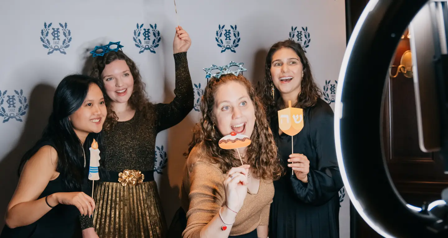 Four women from the congregation smile and hold Hanukkah props in front of a festive backdrop at a Temple Emanu-El event.
