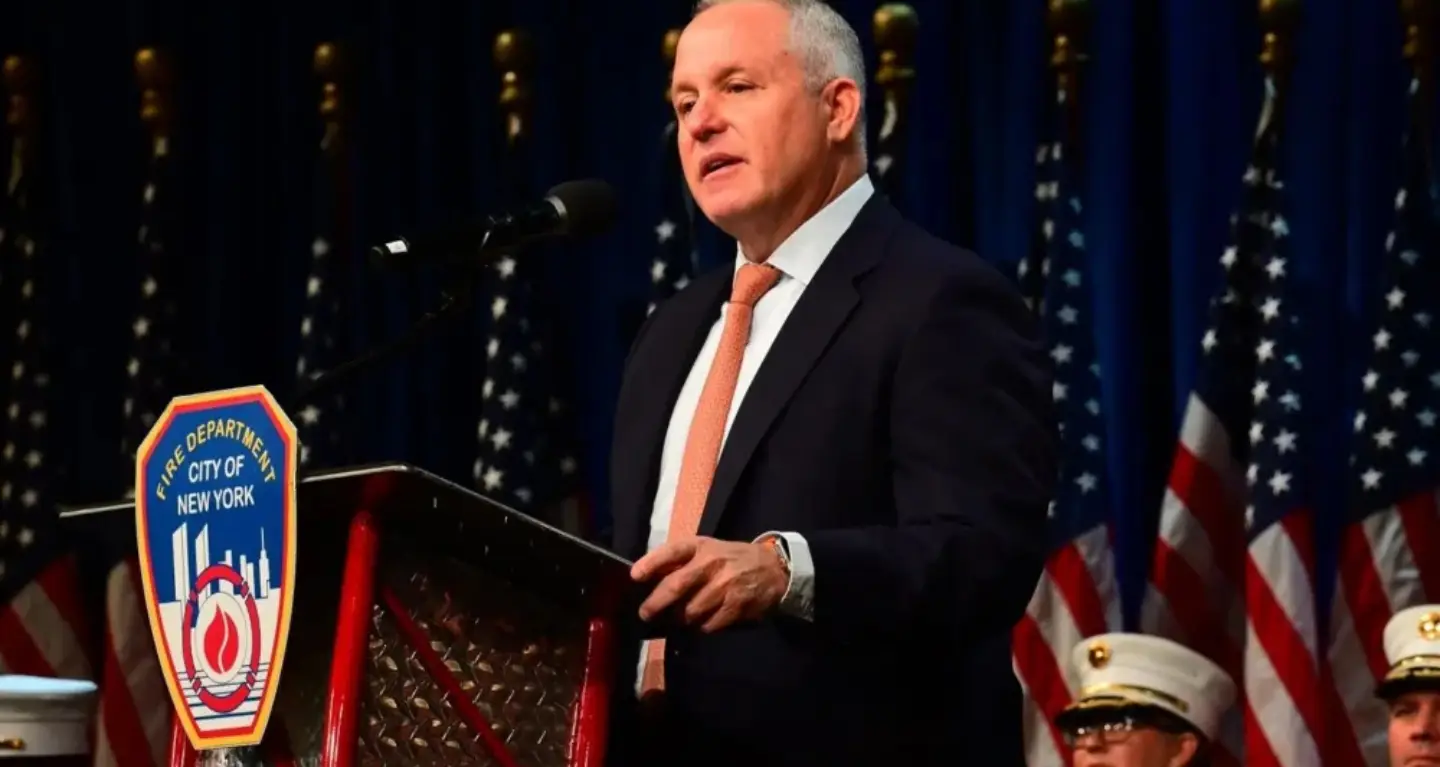 Man in suit speaks at podium with FDNY emblem, flanked by American flags and officials, inside Temple Emanu-El sanctuary.