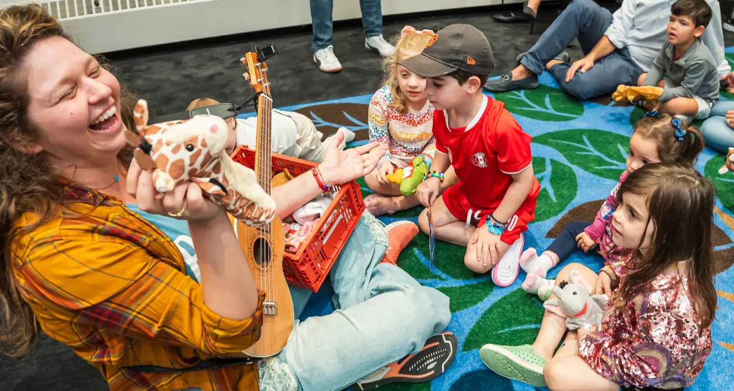 A woman plays ukulele and smiles at children seated in a circle, each holding a stuffed animal, during a Temple Emanu-El program.