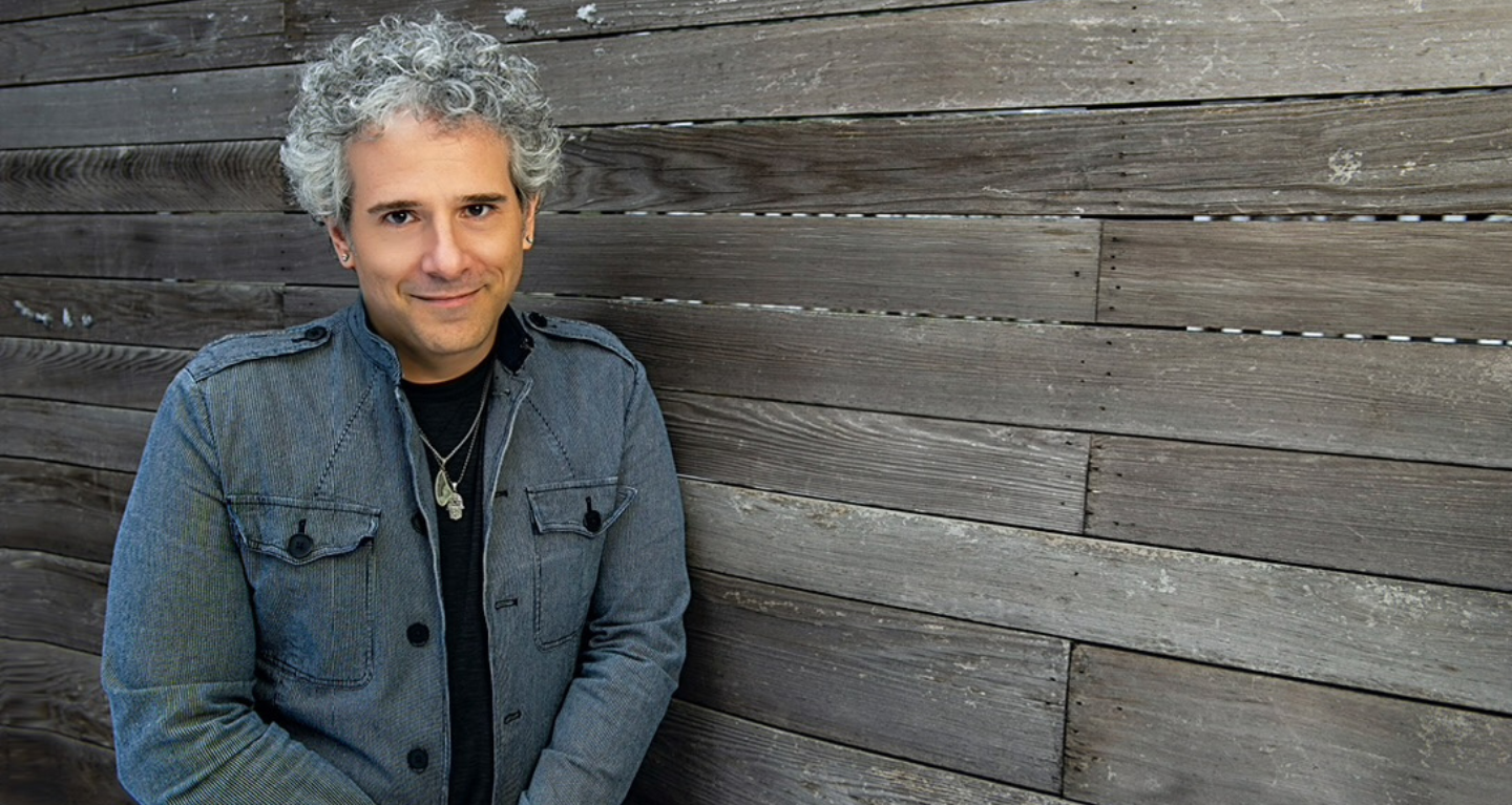 A man with curly gray hair in a gray jacket leans against a wooden wall at Temple Emanu-El, smiling gently.