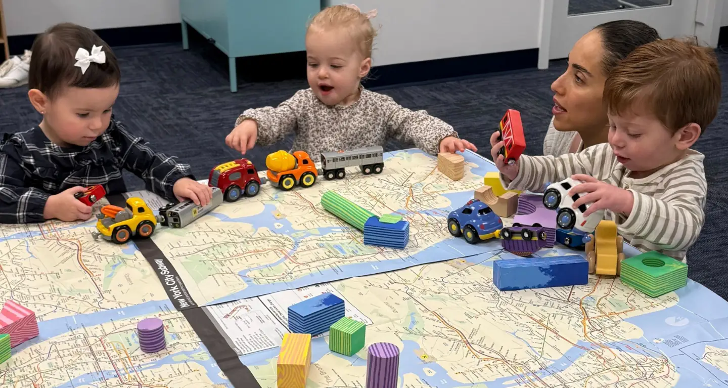 Three toddlers play with toy cars and blocks on a mat as an adult gently supervises, smiling warmly, in a synagogue classroom.