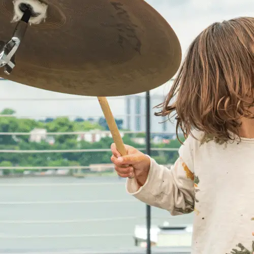 A young child joyfully plays a drum set outdoors near a waterfront railing during a community event at Temple Emanu-El.
