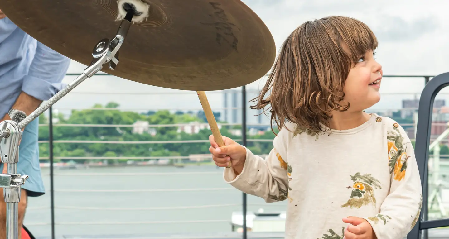 A young child joyfully plays a drum set outdoors near a waterfront railing during a community event at Temple Emanu-El.
