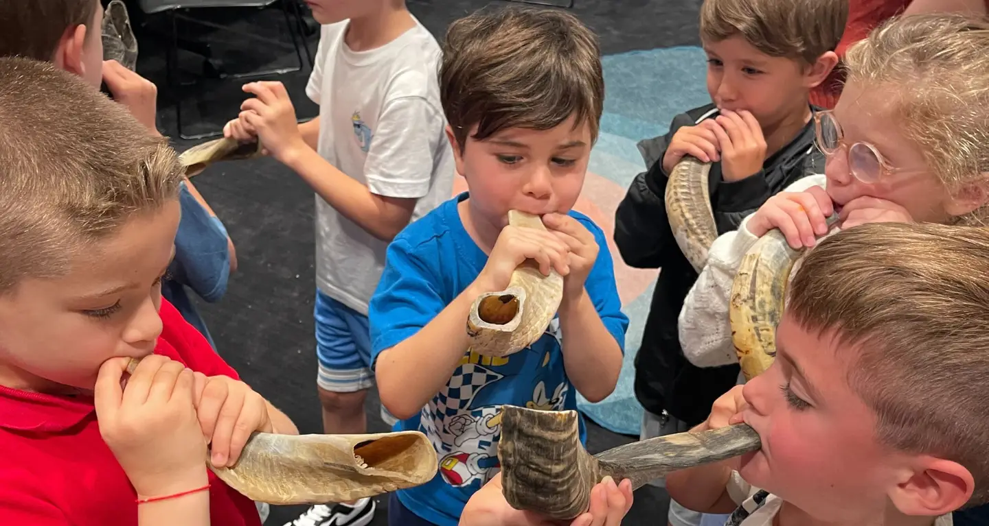 Children stand in a circle, joyfully blowing shofars together in a bright classroom at Temple Emanu-El.