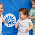 Three children smile in front of a blue banner at Temple Emanu-El, one holding a snack and water bottle.