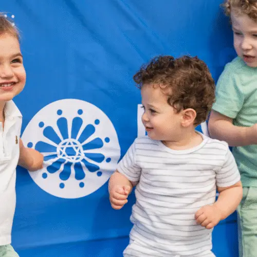 Three children smile in front of a blue banner at Temple Emanu-El, one holding a snack and water bottle.