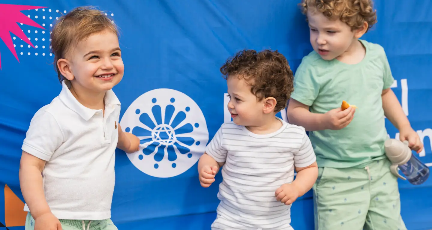 Three children smile in front of a blue banner at Temple Emanu-El, one holding a snack and water bottle.