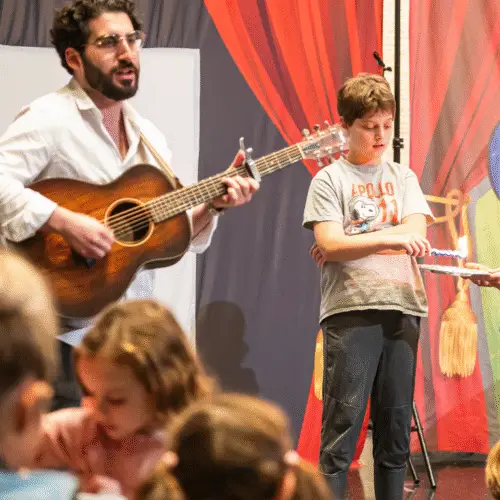 A man plays guitar on stage at Temple Emanu-El while children gather and interact in front of a vibrant backdrop.