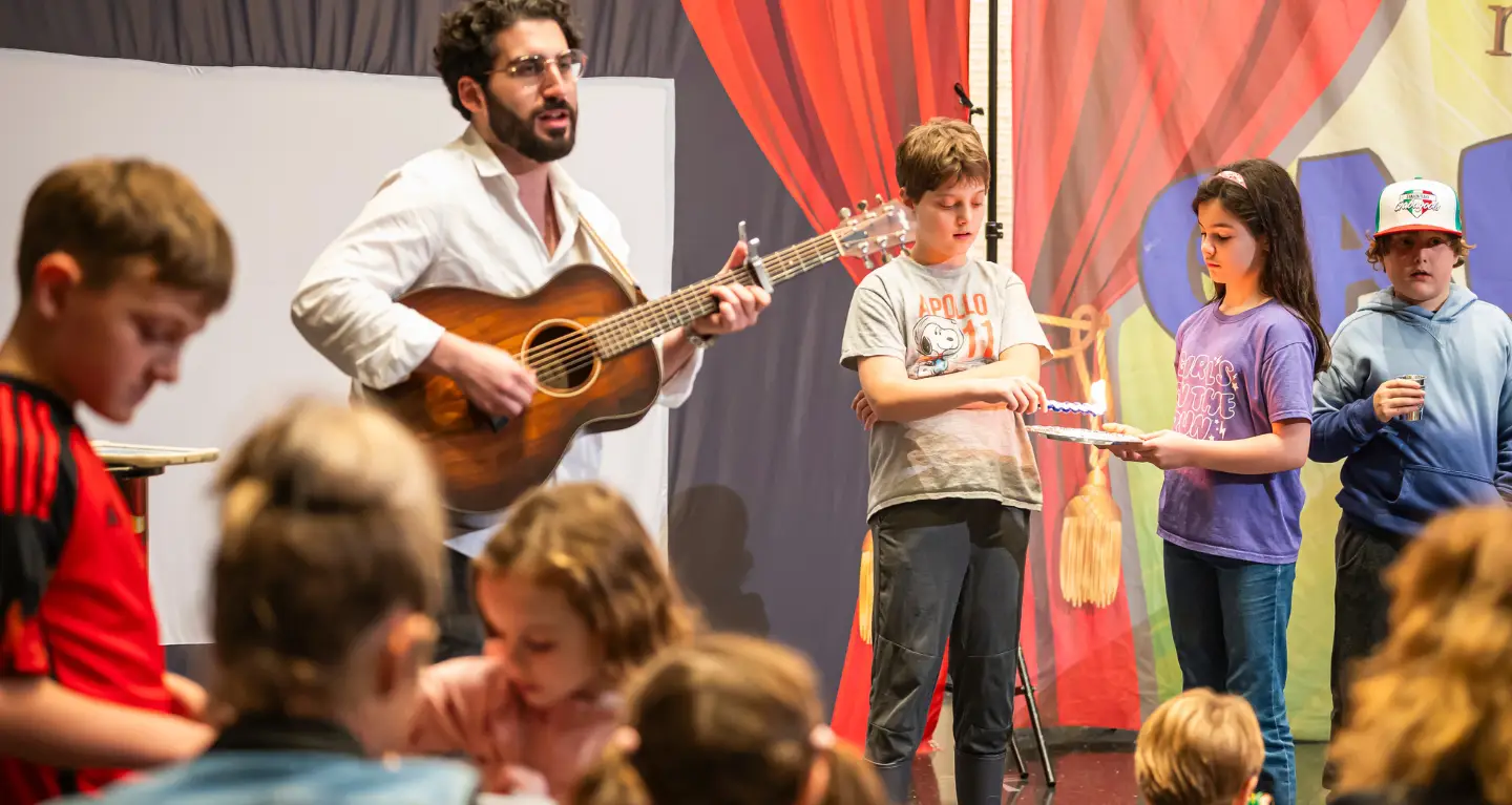 A man plays guitar on stage at Temple Emanu-El while children gather and interact in front of a vibrant backdrop.