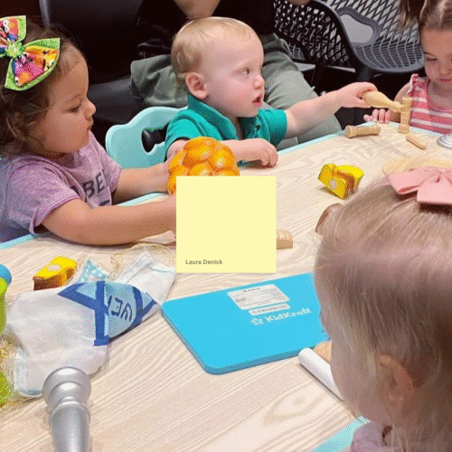 Six young children gather around a table playing with toys, guided by adults in a welcoming Temple Emanu-El classroom setting.