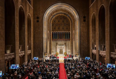 A large congregation gathers inside Temple Emanu-El’s grand sanctuary, with soaring arches and a red carpet down the central aisle.