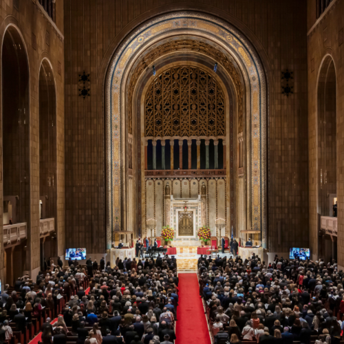 A large congregation gathers inside Temple Emanu-El’s grand sanctuary, with soaring arches and a red carpet down the central aisle.