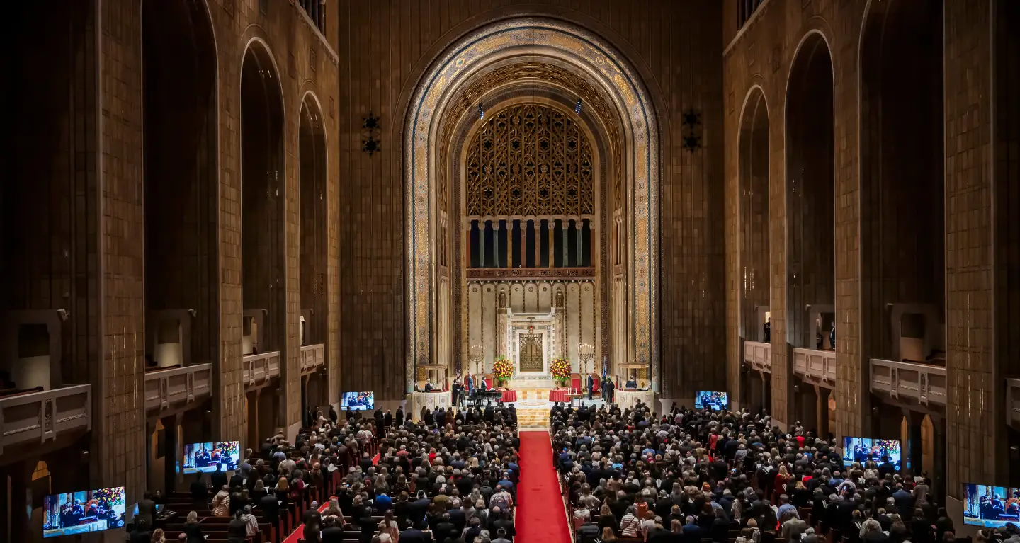 A large congregation gathers inside Temple Emanu-El’s grand sanctuary, with soaring arches and a red carpet down the central aisle.