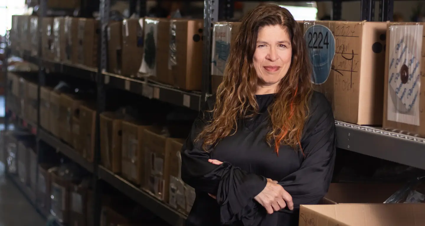 A woman with long hair smiles warmly inside a storeroom, shelves and boxes behind her, reflecting Temple Emanu-El’s community spirit.